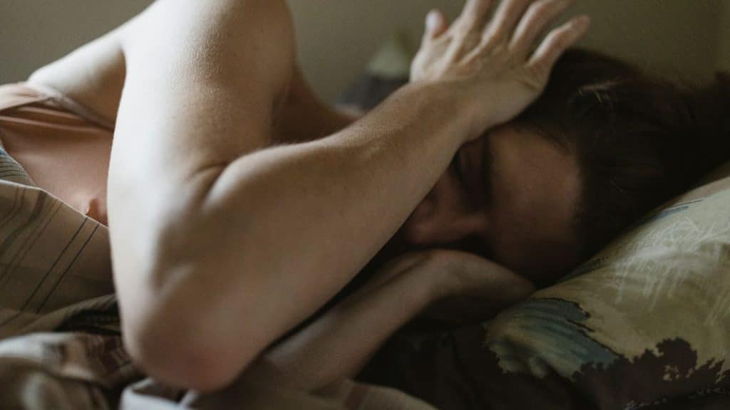 Person lying in bed, partially covered by blankets, covering face with an arm.
