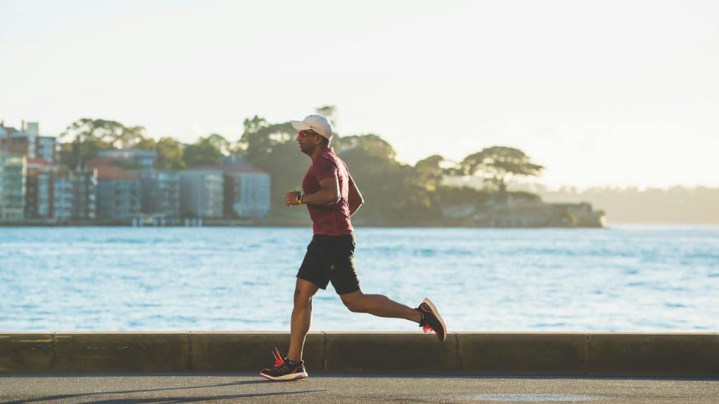 A man in a red shirt and black shorts is running along a paved waterfront area on a sunny day.