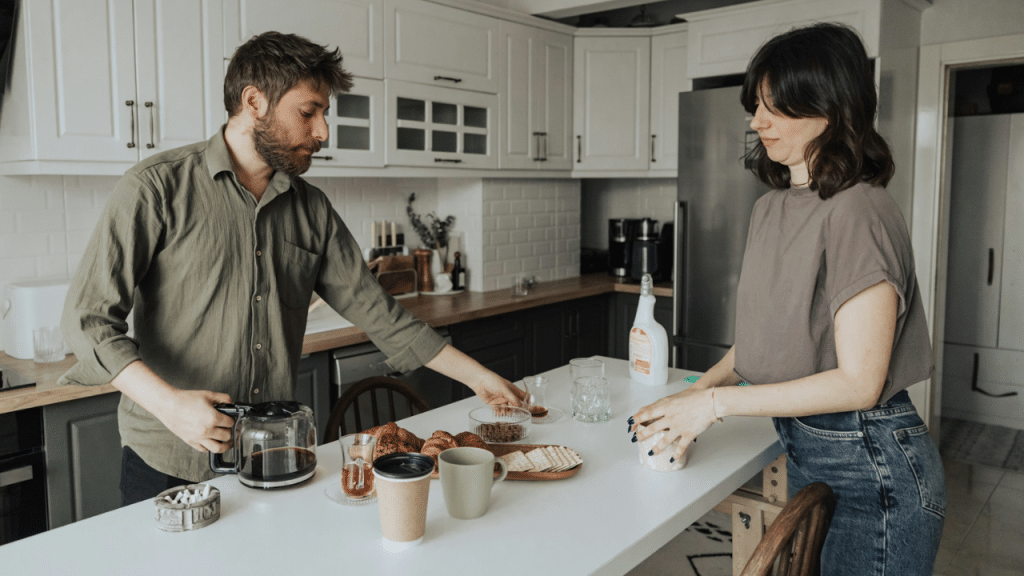 Man in green shirt pouring coffee while woman prepares a kitchen counter meal.