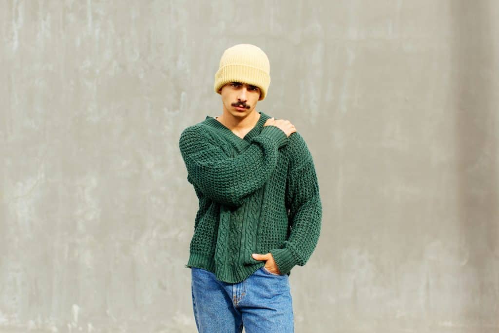 A man standing in front of a wall wearing green sweater.