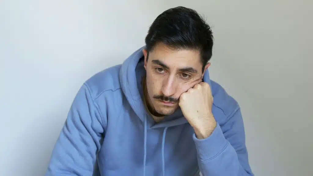 A man with a mustache in a blue hoodie, resting his chin on his hand.