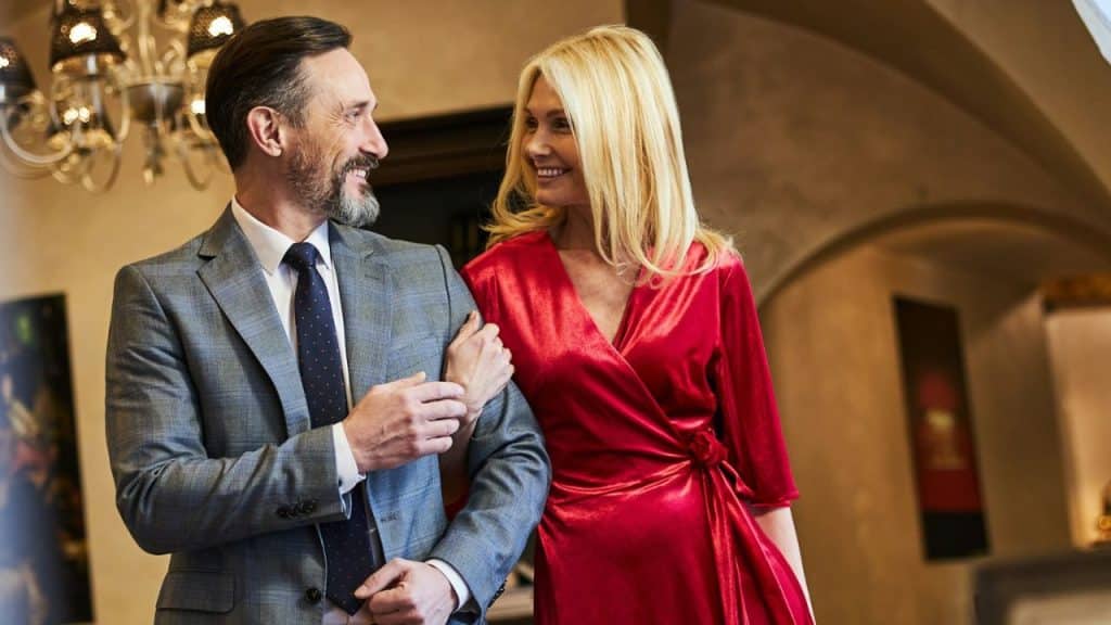 Elegant middle-aged couple smiling while walking arm-in-arm indoors, dressed formally.