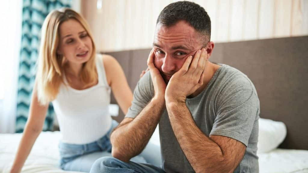 A man sits with his head in his hands, looking distressed, as a woman talks behind him.