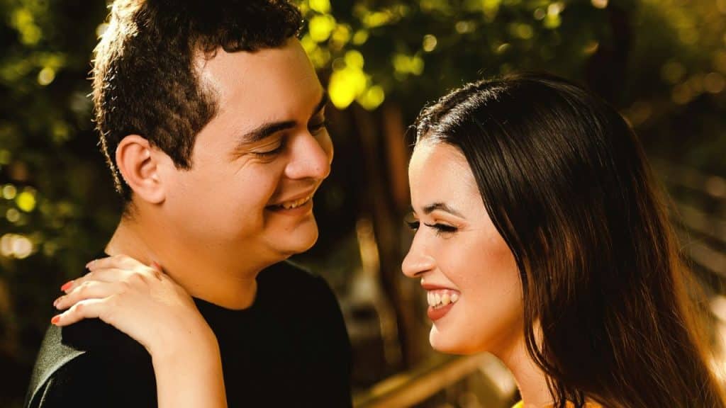 Man and woman smiling at each other in an outdoor setting with green foliage.