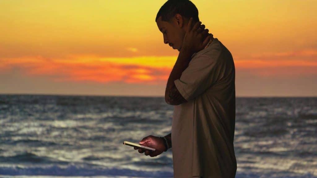 A man standing by the ocean at sunset looking down at his phone with a hand on his neck.