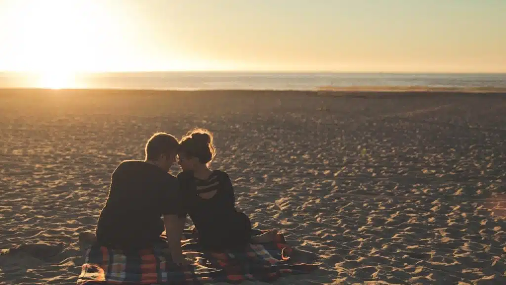 A couple is sitting on a blanket on a beach, facing each other at sunset with their heads touching.