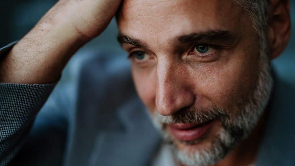 Close-up of a handsome gray-haired man with a beard, looking down with one hand on his head.