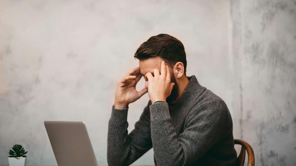 A man sitting at a desk with a laptop, holding his head in stress.