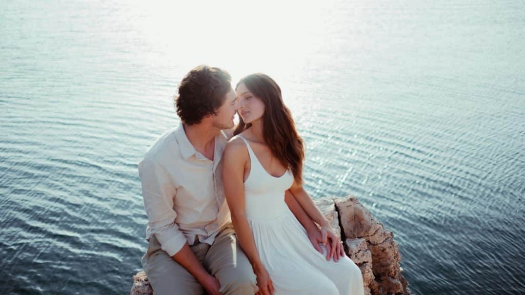 A couple sits on a rocky outcrop over calm water, facing each other.