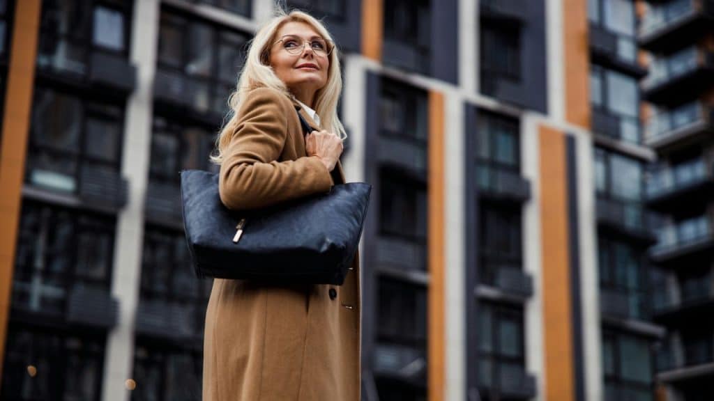 A fashionable blonde woman looks up confidently while holding a purse outdoors.