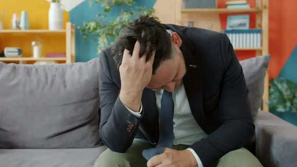 Man in a suit jacket and tie sitting on a couch, head down, hand in his hair.