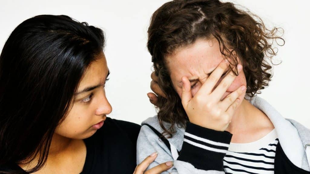 Two young women, one comforting the other who is covering her face.