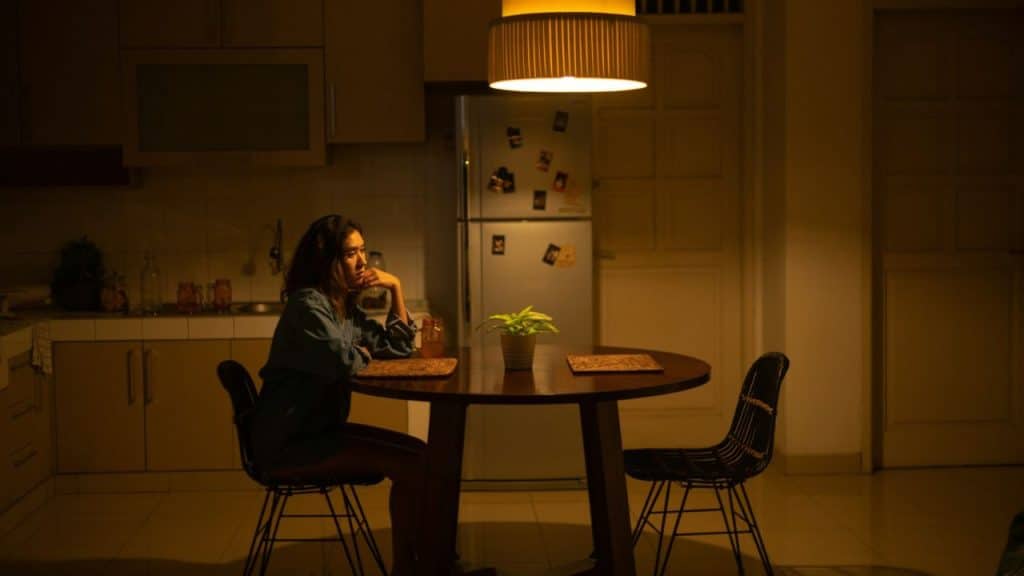 A woman sits alone at a dimly lit kitchen table, resting her head on her hand.