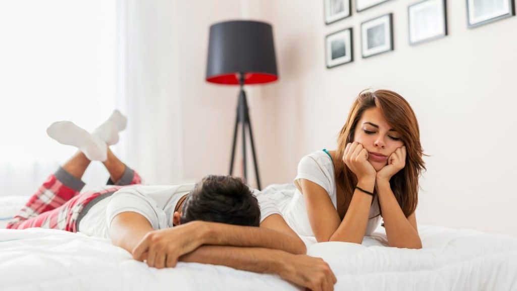 A man and woman lie on a bed, the woman resting her chin on her hands, looking bored.