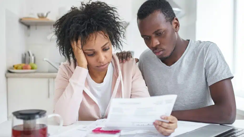 A woman looks stressed holding her head while a man reads a paper with his arm around her.