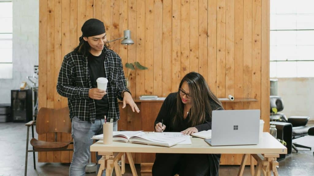 A man in a bandana talking to a woman writing on papers at a wooden desk indoors.