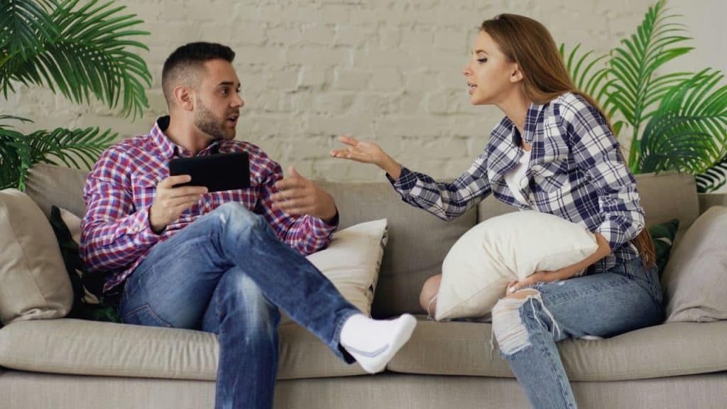 A couple argues on a couch; the woman is gesturing while the man holds a tablet.