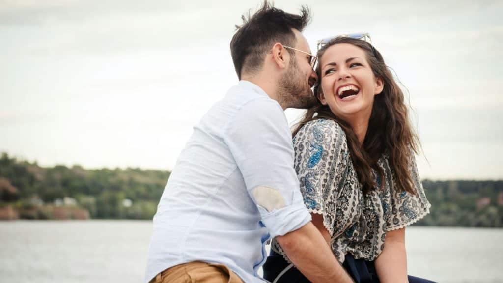 Smiling man and woman outdoors, man whispering into woman's ear, laughing happily.`