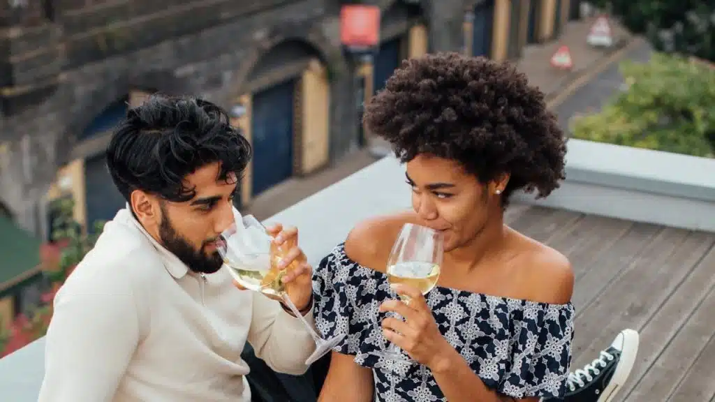 A couple sits on a wooden deck or rooftop patio, sipping white wine and enjoying a picnic of grapes and roses.