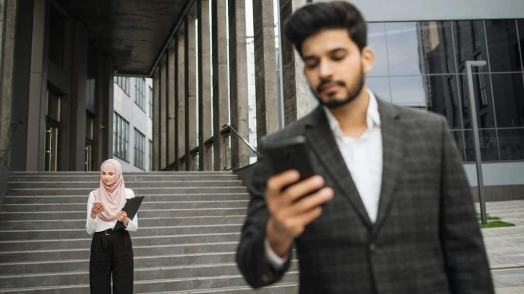 A man in the forefront looking at his phone while a woman wearing a scarf looks at her phone in the background.