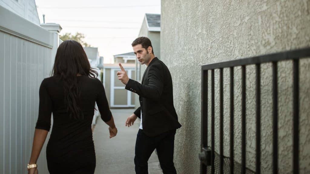 A man in a black jacket is arguing with a woman walking away from him in a black dress, gesturing with his hand.