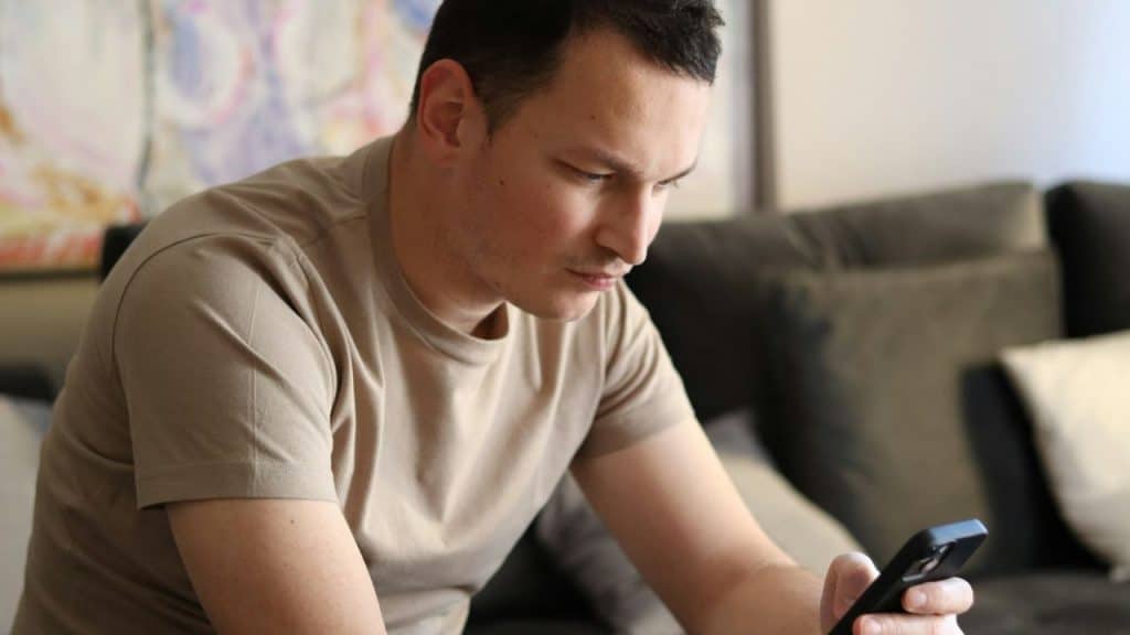 Man in a tan shirt sitting on a couch, looking intently at his black smartphone.