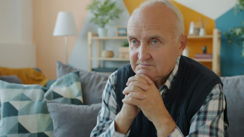 Close-up of a serious elderly man sitting on a couch with hands clasped.