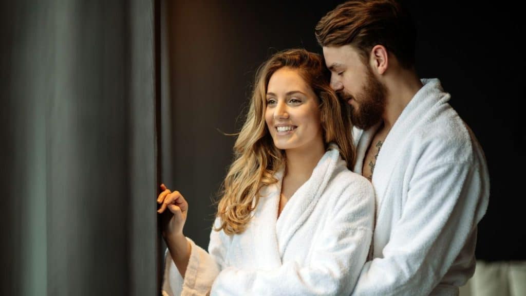 Man and woman in white robes embrace, looking out a window in soft light.