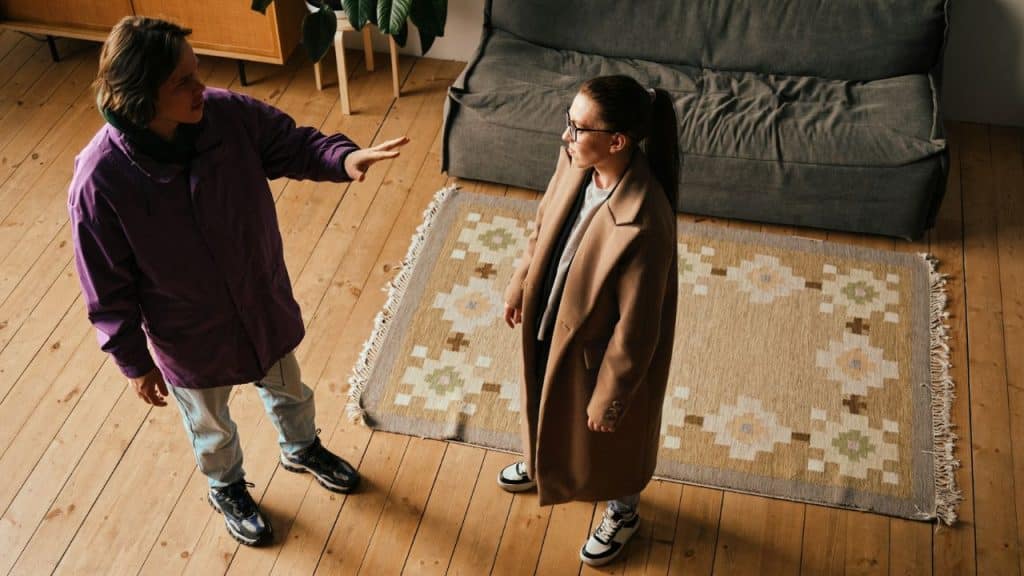 A man in a purple jacket talks to a woman in a tan coat on a wooden floor.