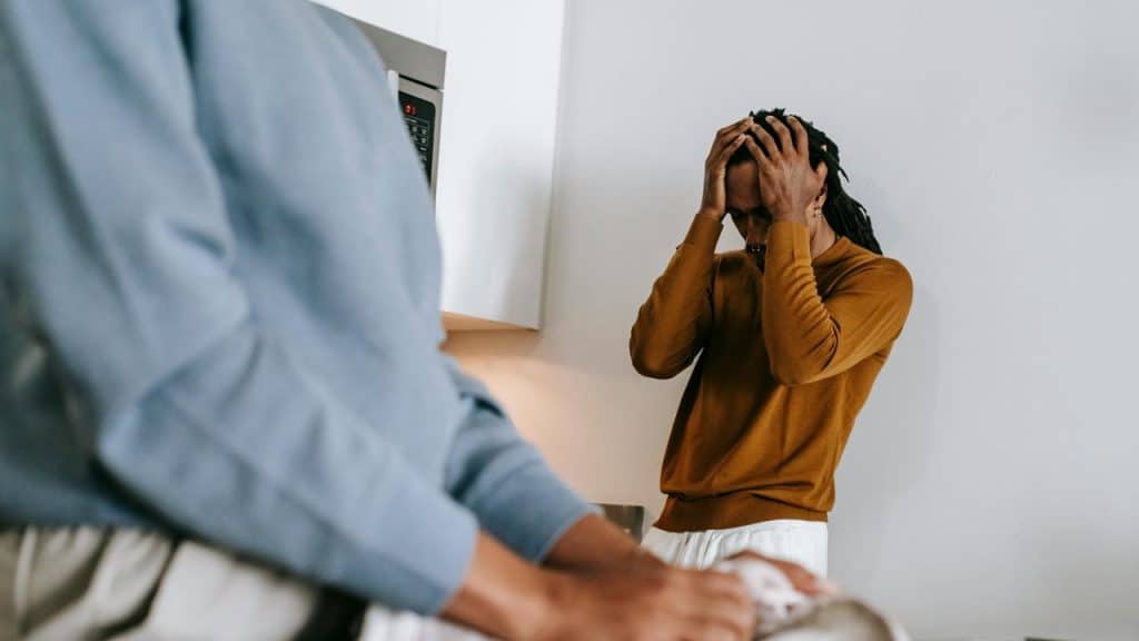 A man standing in a kitchen with his hands on his head while another person faces him.