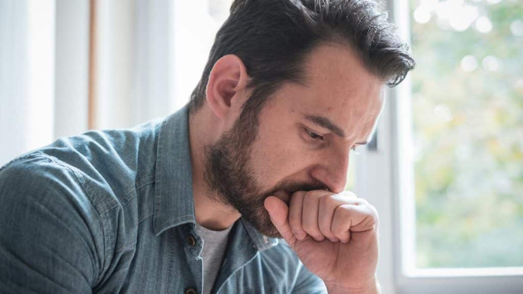 A close-up shot of a worried or distressed man with a beard, holding his fist to his mouth.