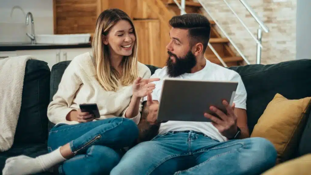 Smiling couple relaxing on a dark sofa, looking at a digital tablet.
