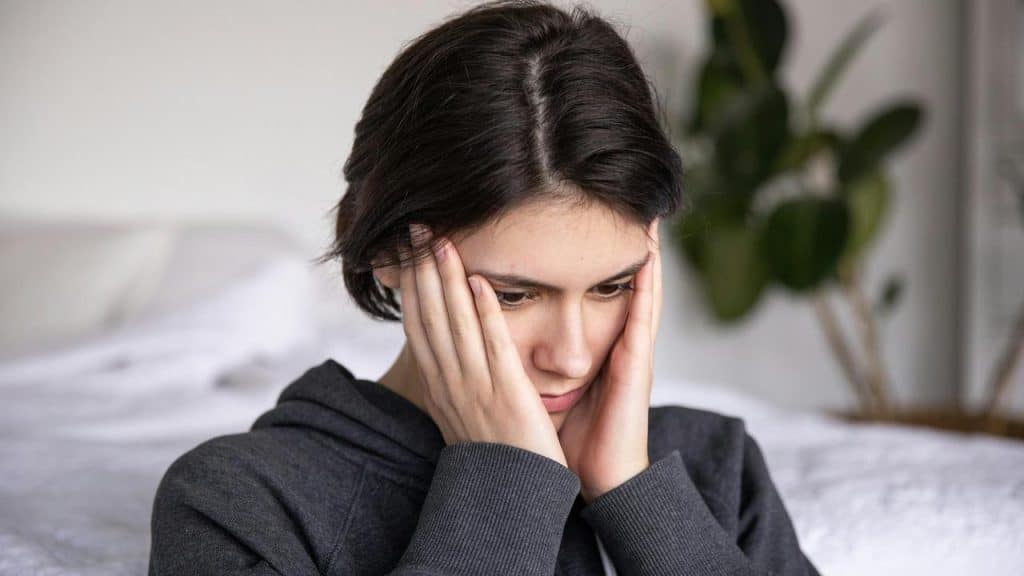 A woman sitting indoors with her hands on her face looking down.