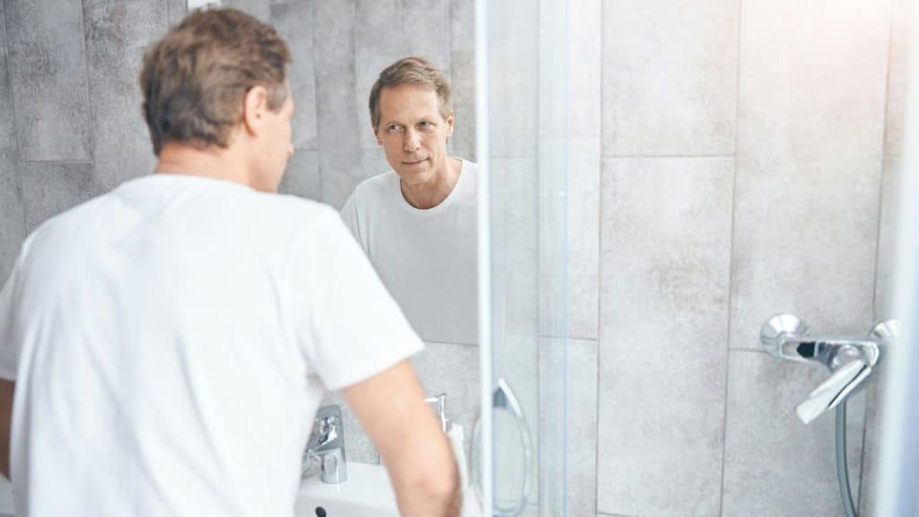 Man in white shirt looking at his reflection in a bathroom mirror.