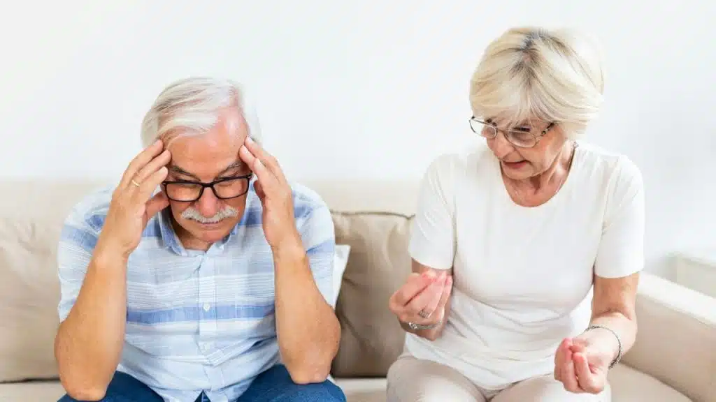Elderly man holds his head in his hands next to a talking elderly woman.
