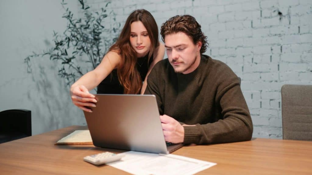 A man and woman look intently at a laptop screen on a wooden desk.