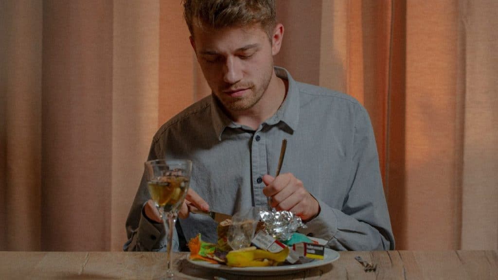 Man eating from a plate of garbage with a fork and knife, with a glass of wine.