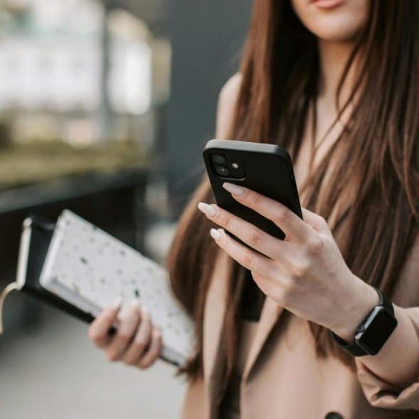 A woman holding a notebook checks her phone while standing outdoors.