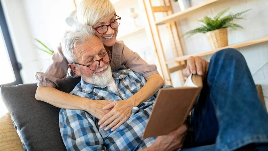 A smiling woman hugging a man while he reads a book on the couch.