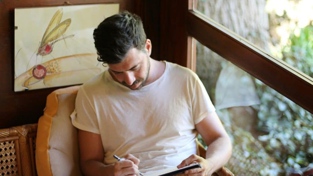 A man sitting by a window writing in a notebook.