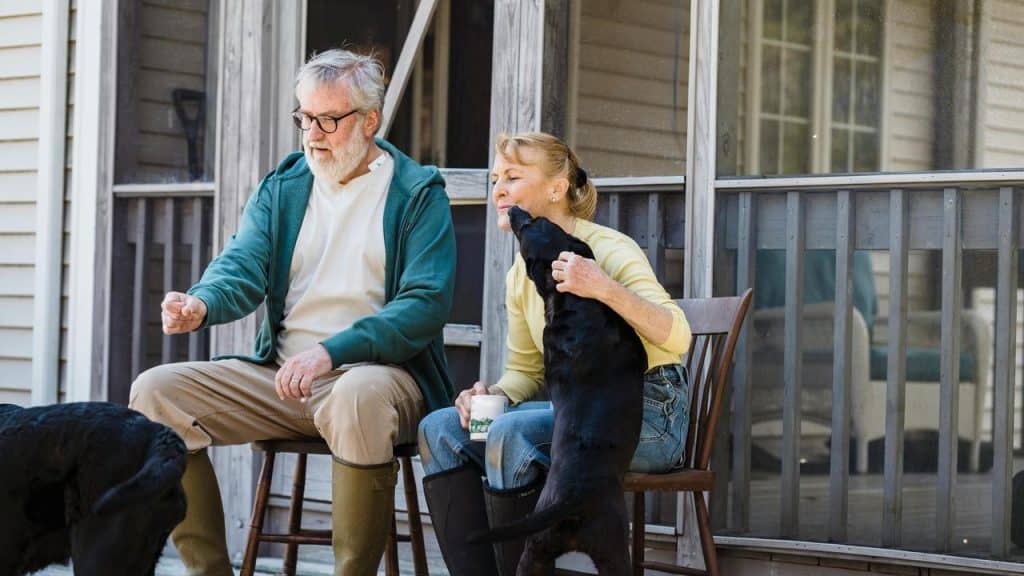 A couple sitting on a porch while a black dog stands up affectionately beside them.