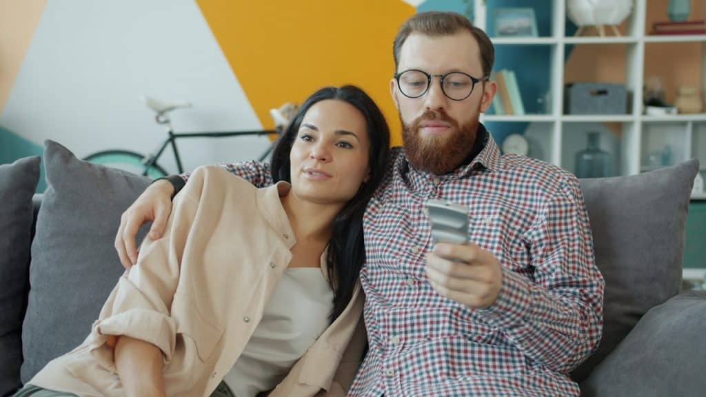 A bearded man wearing glasses using his phone and a woman leaning on him with his arm around her shoulder as they sit on a couch.