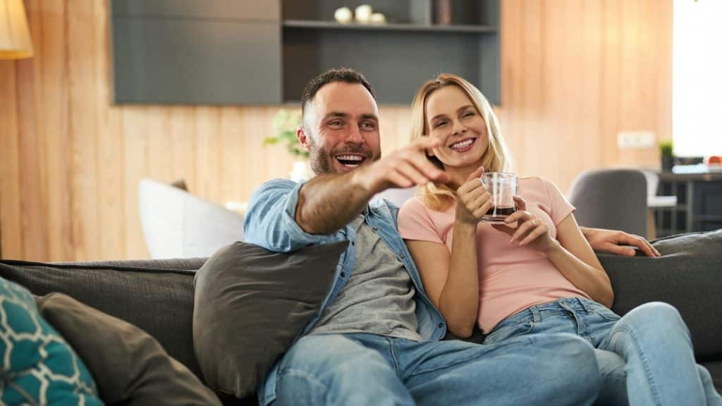 A couple laughing while sitting on the couch and the man points towards the TV.