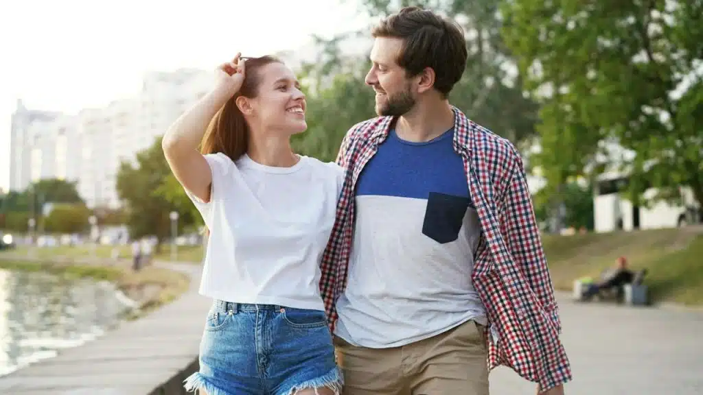 A man and woman walk along a waterfront path, smiling at each other and arm-in-arm.