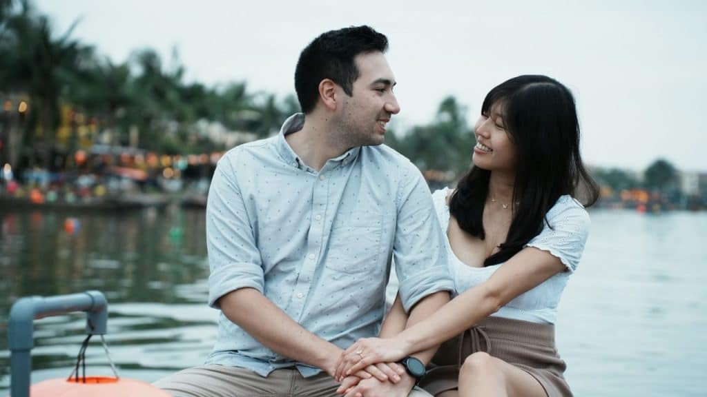 A man and woman sit near water, holding hands and smiling while looking at each other.