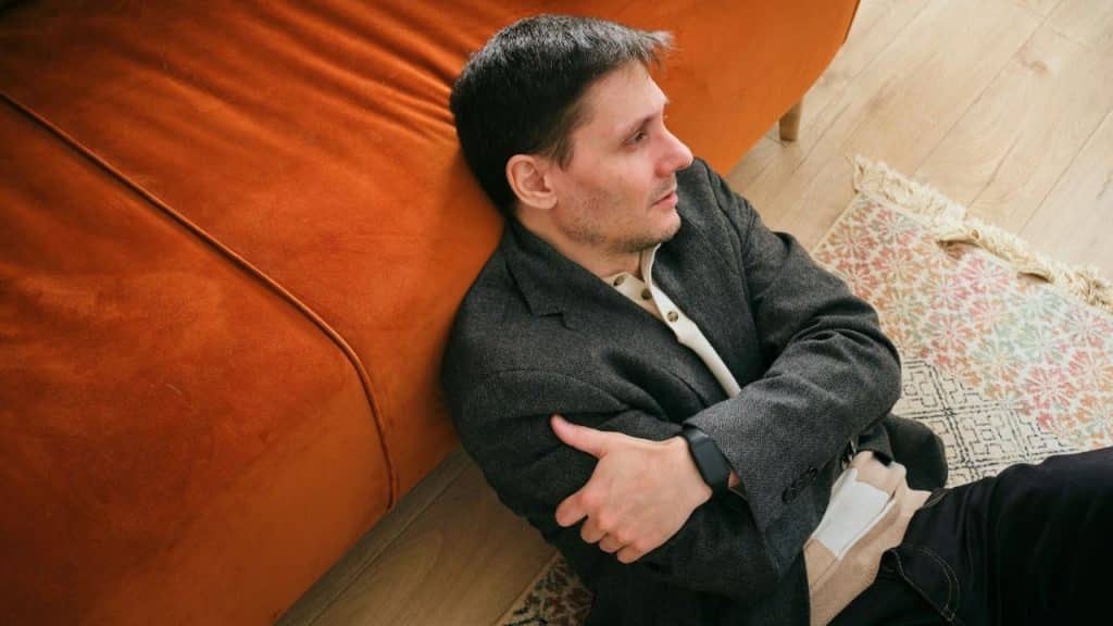 A man in a dark jacket sits on the floor, leaning against an orange sofa.