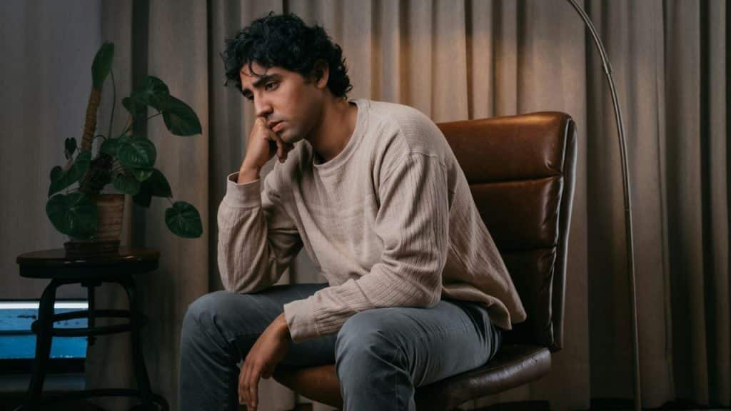 A man with dark, curly hair sits in a brown leather chair, resting his chin on his hand.
