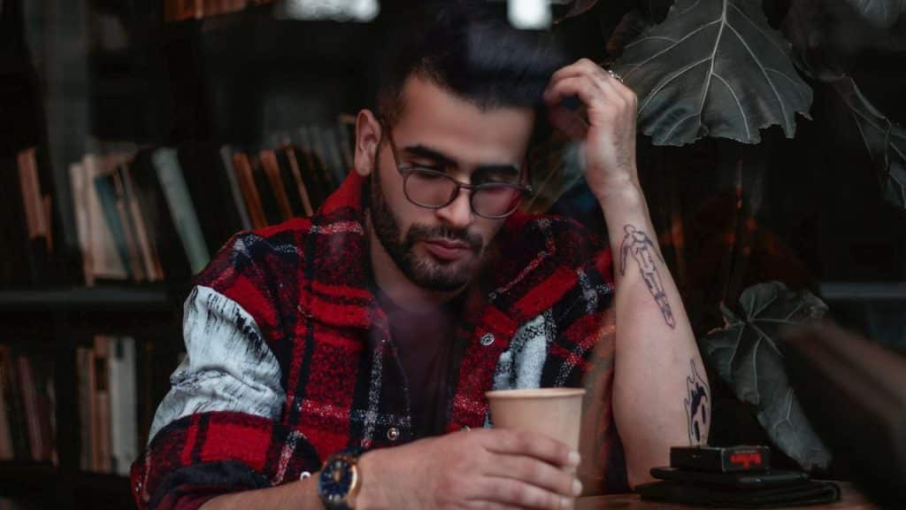 A man in a red plaid jacket and glasses sits at a table with a cup.