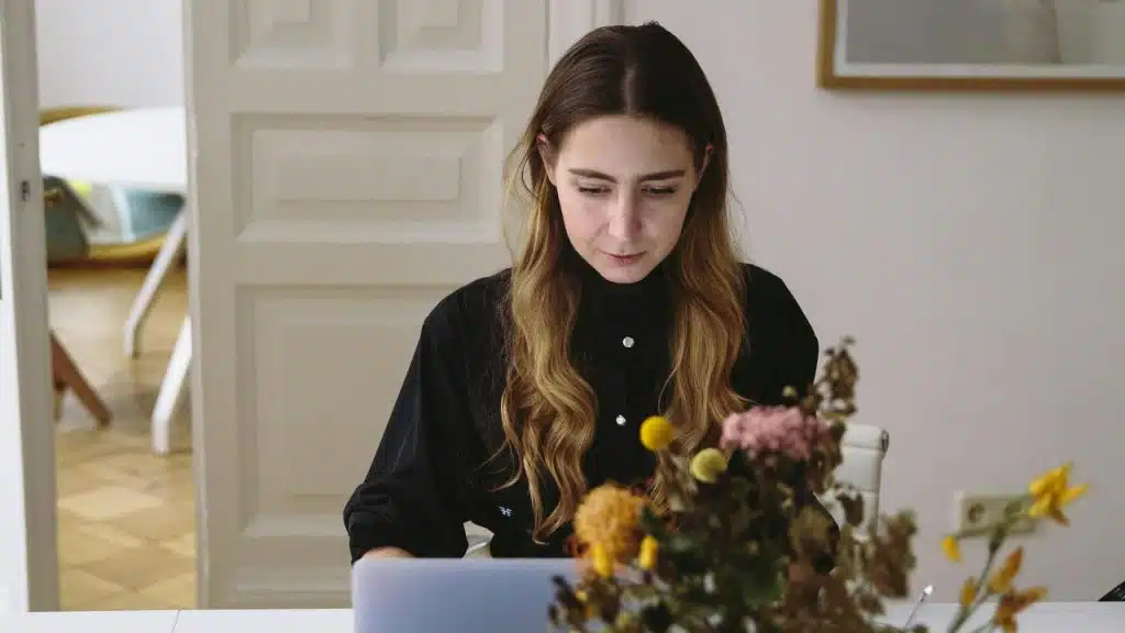 A woman working on a laptop at a desk with flowers in the foreground.
