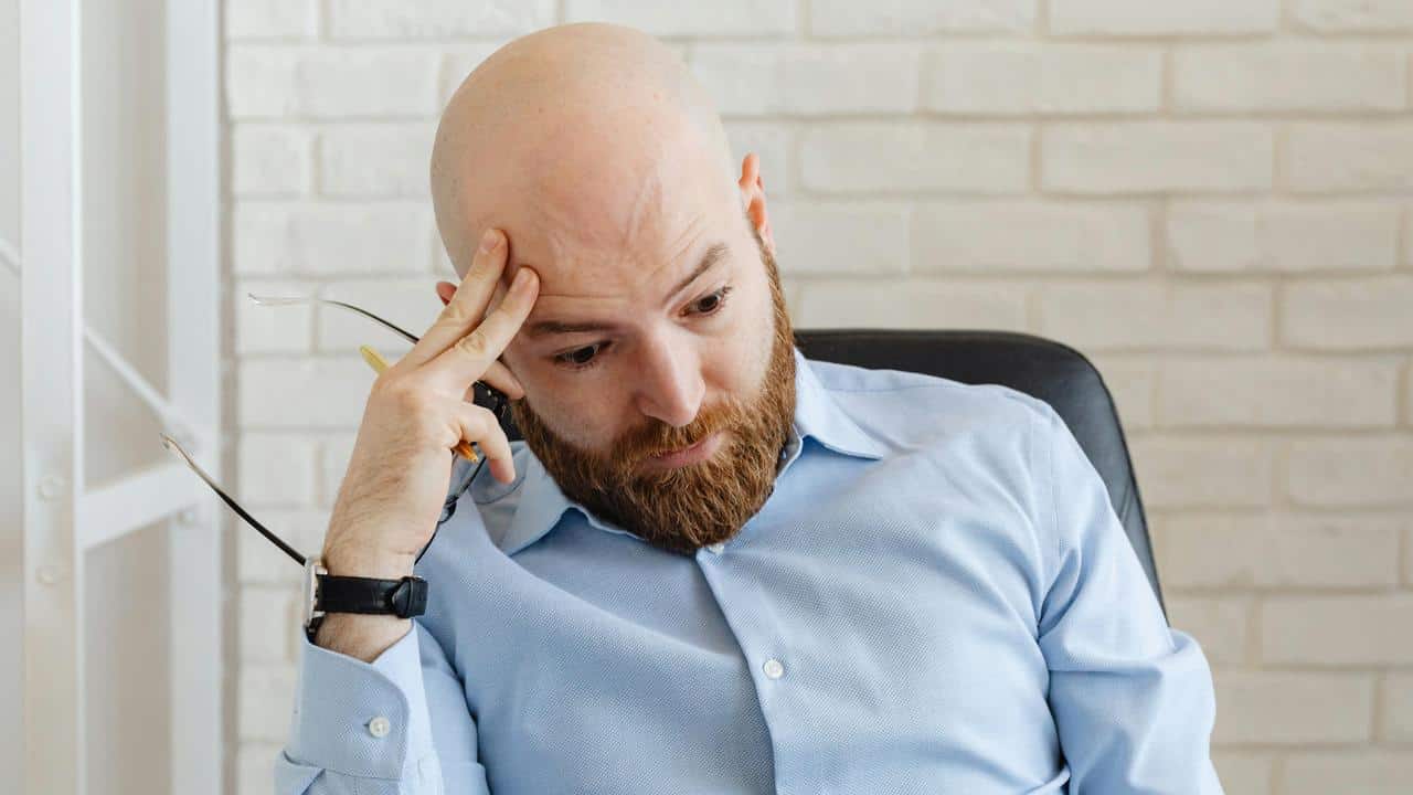 A bald man with a beard looks stressed while holding his forehead.